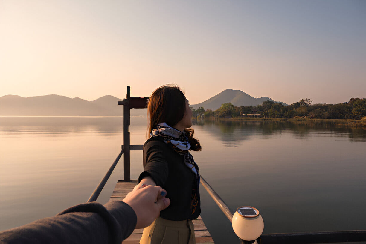 Young couple holding hands on a wooden pier, watching a serene sunrise over a mountain lake in rural Kashmir, capturing the romance of Kashmir Nirvana’s Luxury Kashmir Honeymoon Package.